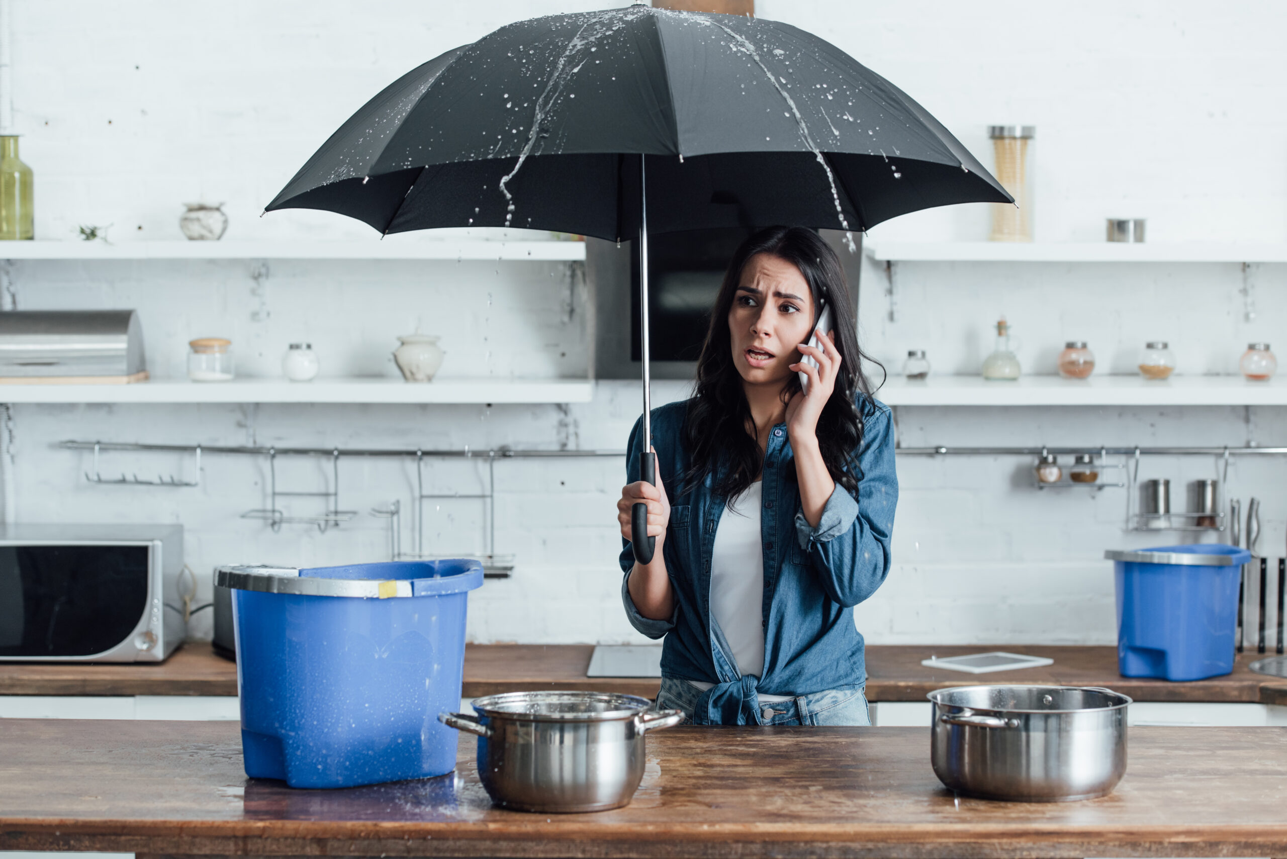 Upset woman standing under umbrella in kitchen and calling a storm damage claim attorney